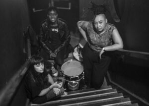 black and white image, three band members pose on a staircase, with drums in the middle of them